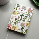 A 2027 daily planner with a white cover decorated with a vintage-style botanical floral pattern, shown on a gray tabletop with a white ceramic mug and a green leaf-shaped plate in the background.