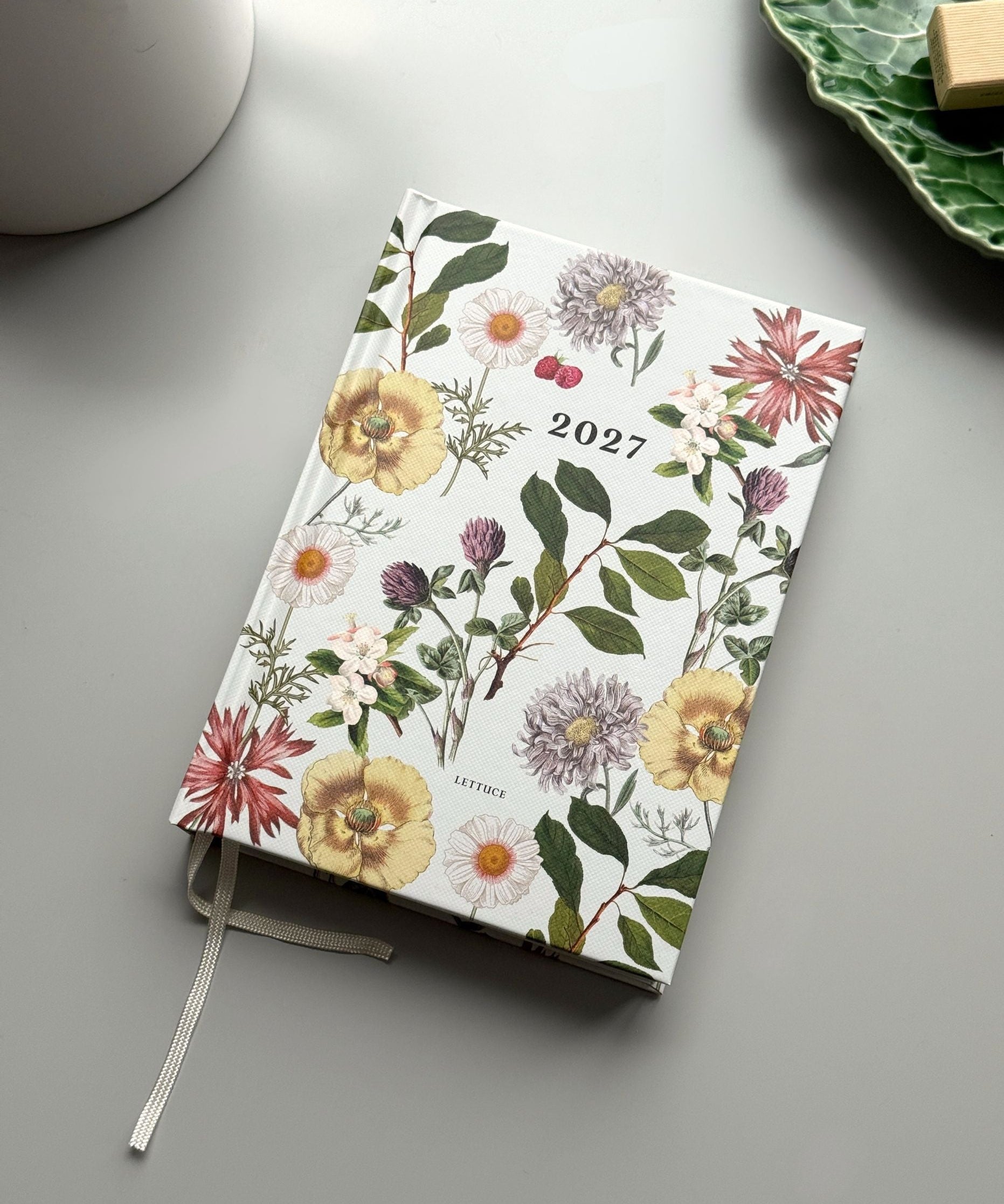 A 2027 daily planner with a white cover decorated with a vintage-style botanical floral pattern, shown on a gray tabletop with a white ceramic mug and a green leaf-shaped plate in the background.