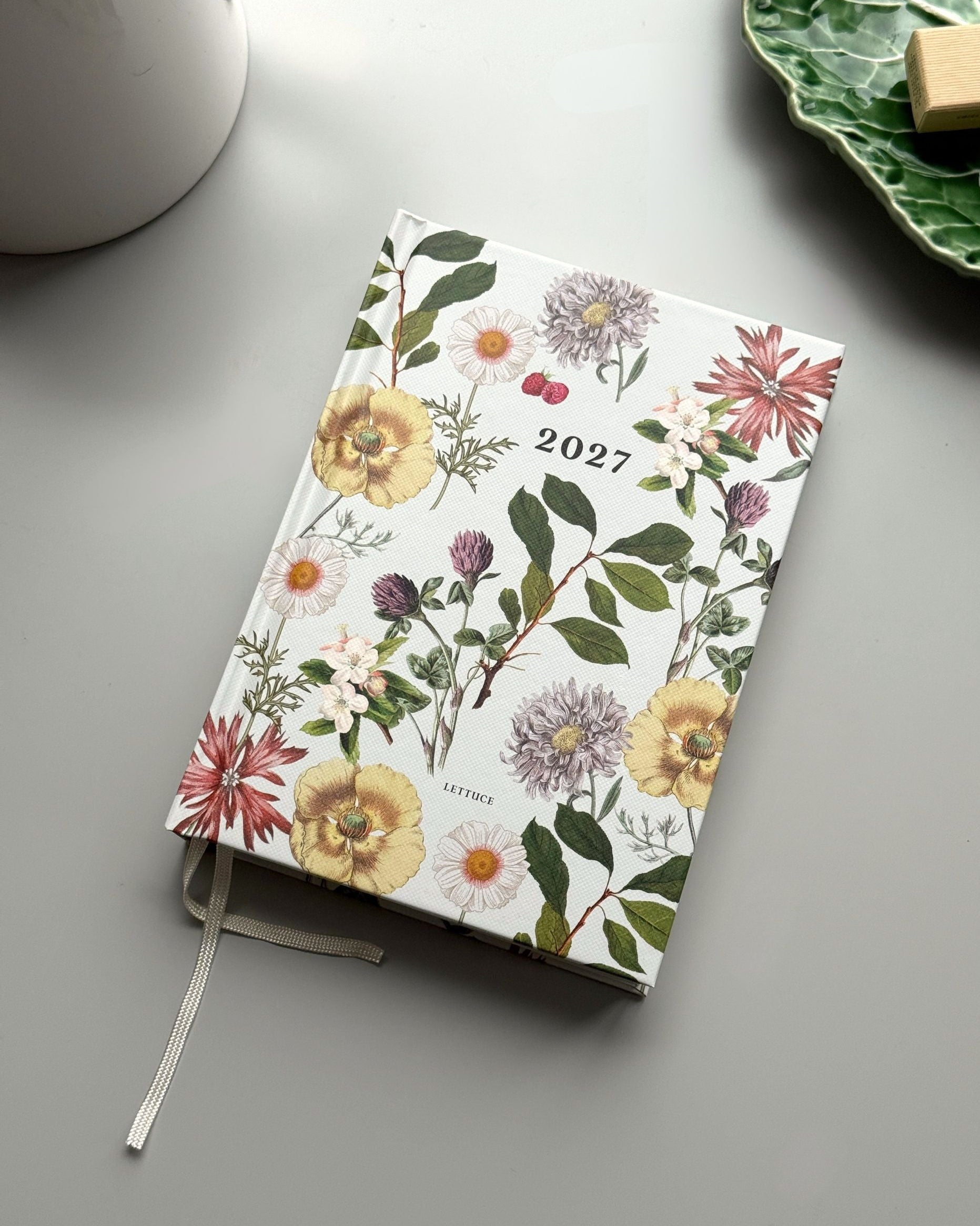 A 2027 daily planner with a white cover decorated with a vintage-style botanical floral pattern, shown on a gray tabletop with a white ceramic mug and a green leaf-shaped plate in the background.