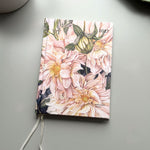 A hardcover planner or journal with a floral cover design featuring pink dahlia flowers and green leaves, shown on a light gray surface with a white ribbon bookmark.