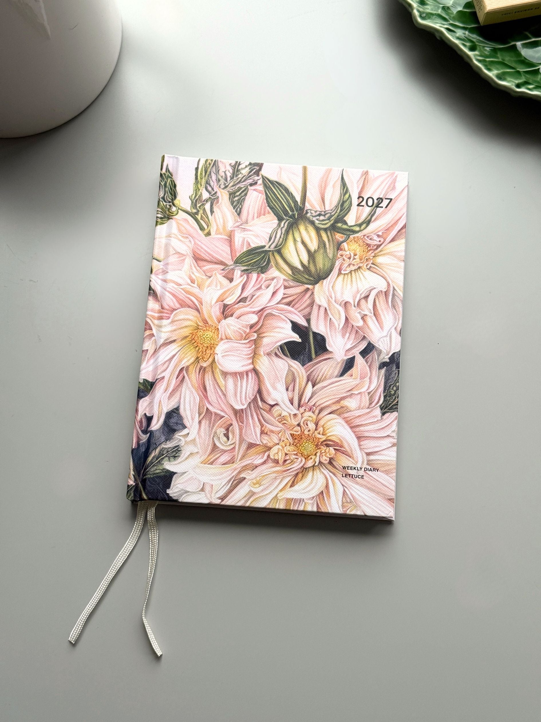 A hardcover planner or journal with a floral cover design featuring pink dahlia flowers and green leaves, shown on a light gray surface with a white ribbon bookmark.