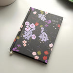 A 2027 weekly planner with a black cover decorated with a colourful floral pattern, resting on a light grey surface with a white ceramic mug partially visible in the top left.