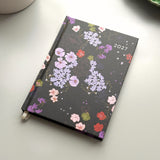 A 2027 weekly planner with a black cover decorated with a colourful floral pattern, resting on a light grey surface with a white ceramic mug partially visible in the top left.