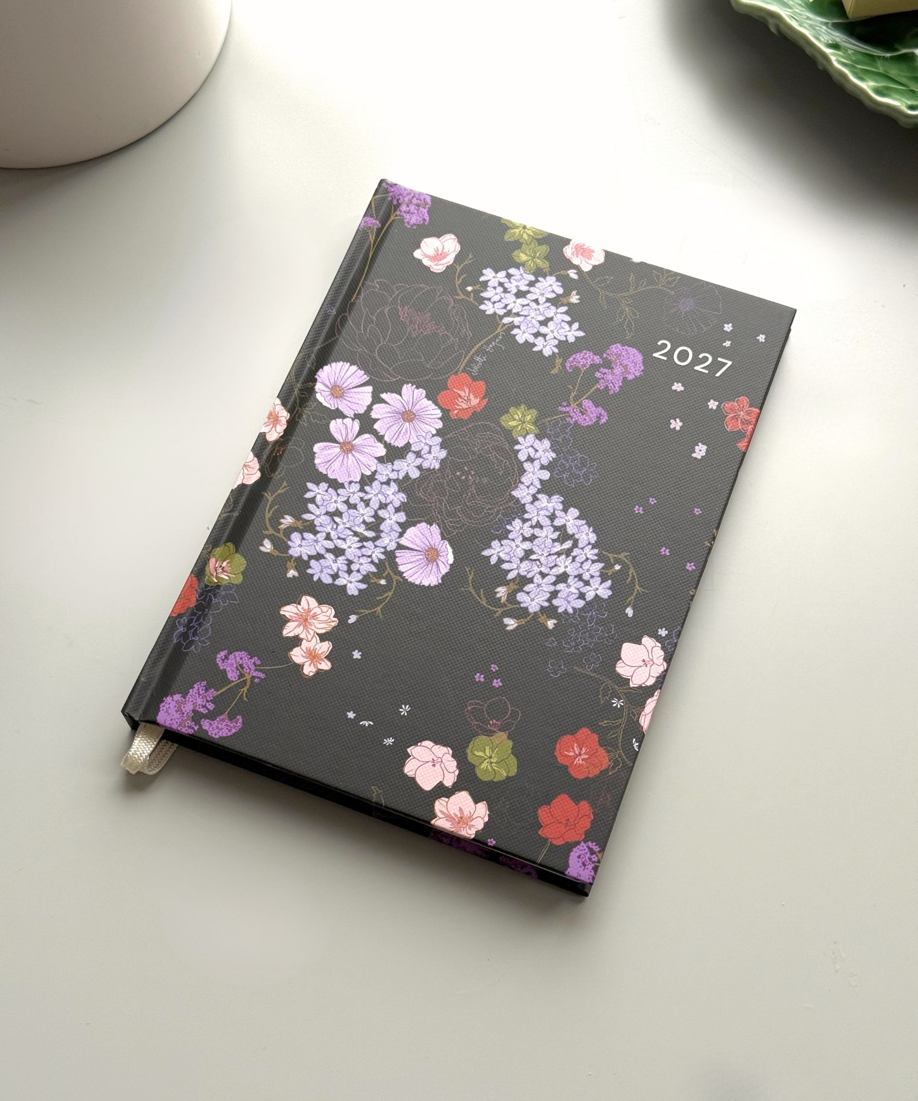 A 2027 weekly planner with a black cover decorated with a colourful floral pattern, resting on a light grey surface with a white ceramic mug partially visible in the top left.