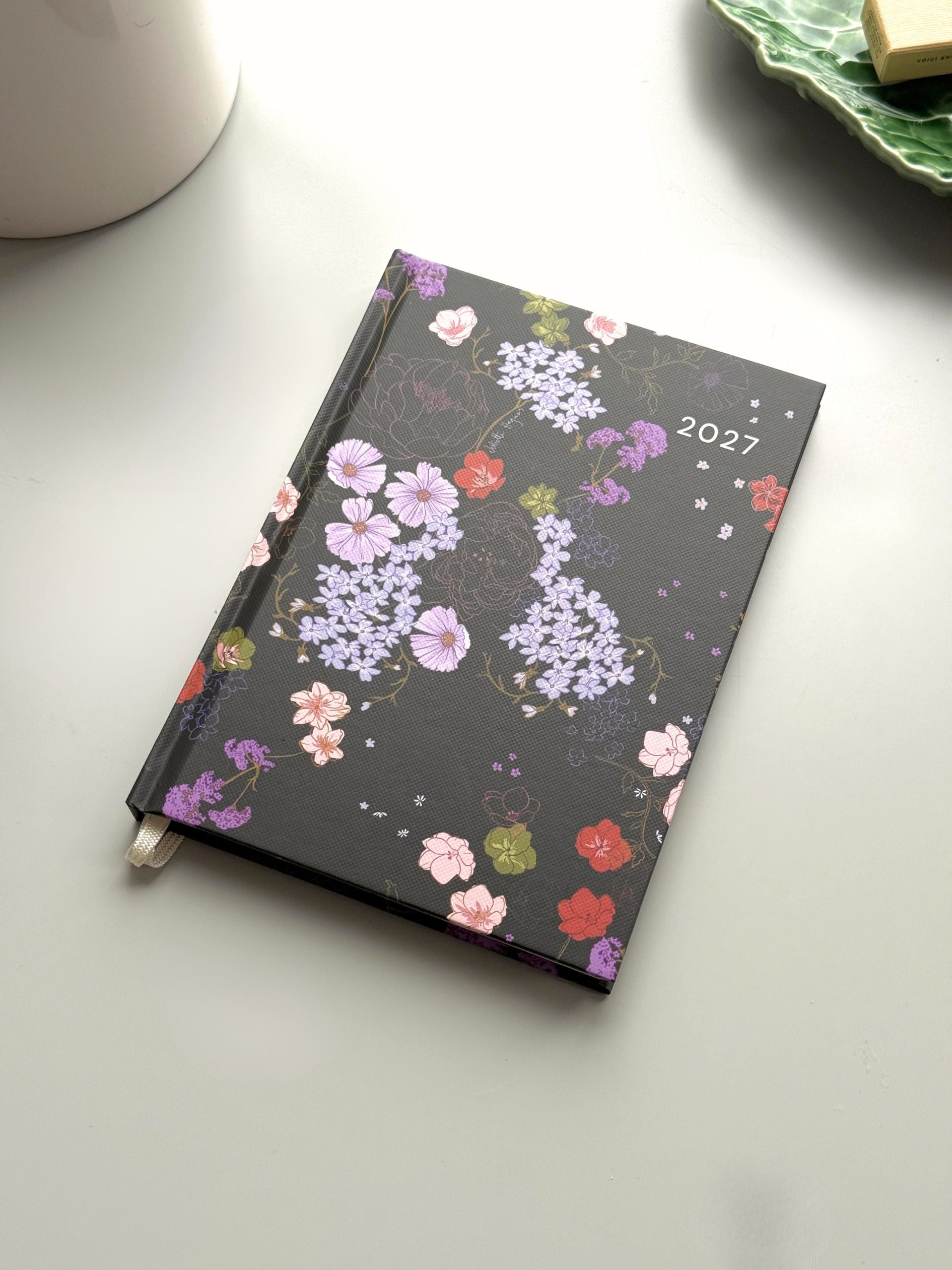 A 2027 weekly planner with a black cover decorated with a colourful floral pattern, resting on a light grey surface with a white ceramic mug partially visible in the top left.